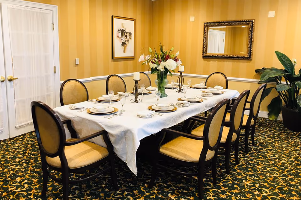 Formal dining room with a long table set for eight, upholstered chairs, a floral centerpiece, and a framed mirror on a striped wall.