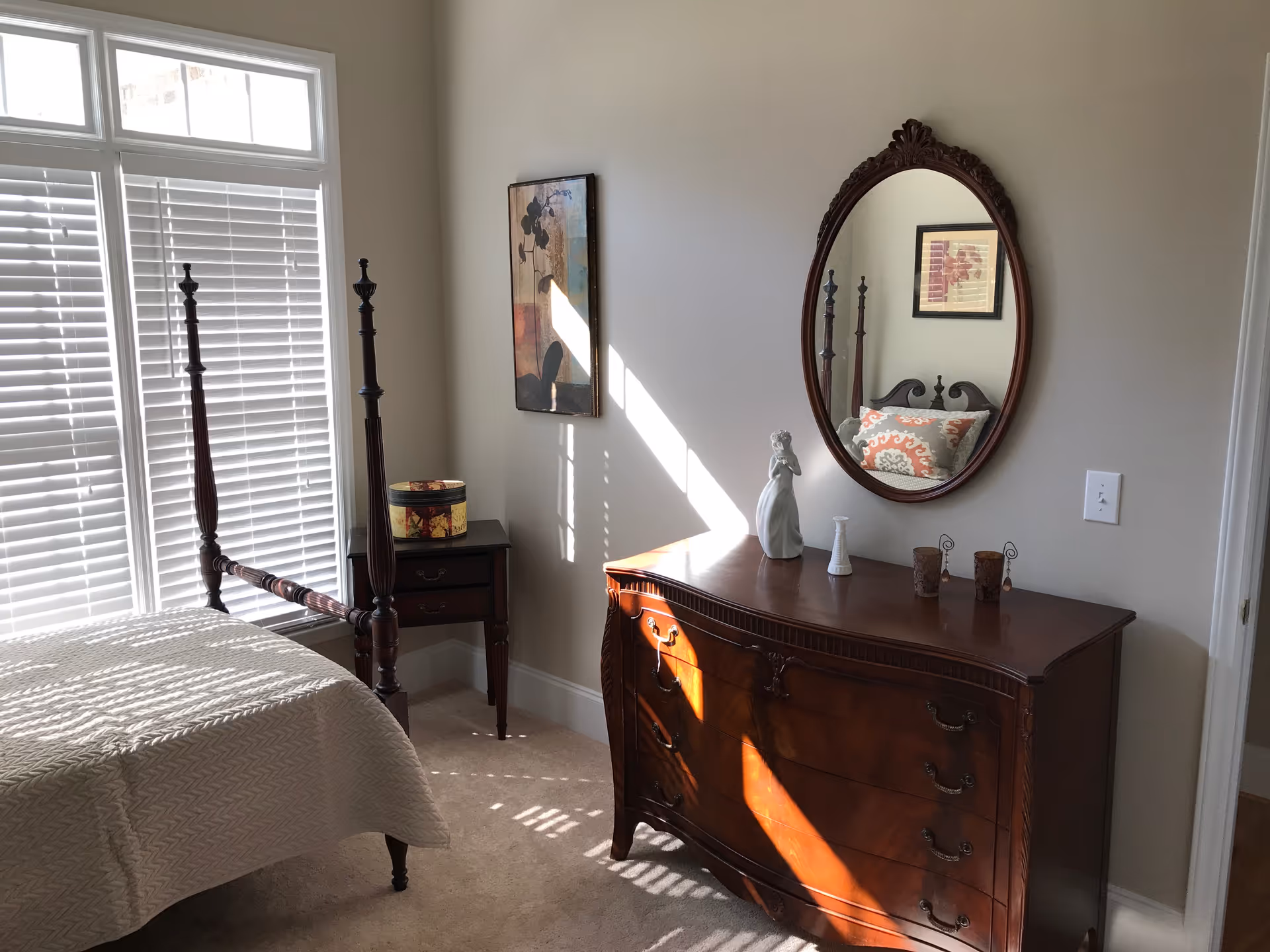 A bedroom with a four-poster bed covered with a beige quilt, a wooden nightstand with a decorative box, and a wooden dresser with an oval mirror above it. The dresser has decorative items including a figurine and small vases. Sunlight streams through white blinds on a large window, casting shadows on the carpeted floor and furniture. A framed artwork hangs on the wall between the window and the dresser.
