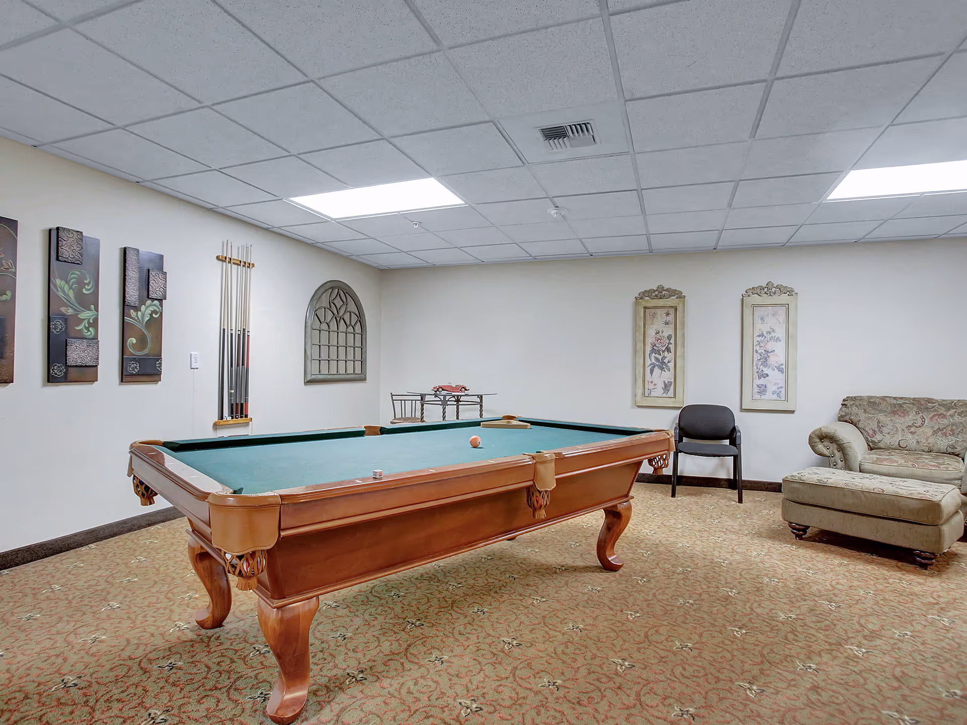A recreational room with a pool table in the center, a wall-mounted rack holding pool cues, decorative wall art, a single chair, and a patterned sofa with an ottoman. The room has a carpeted floor and a drop ceiling with fluorescent lights.