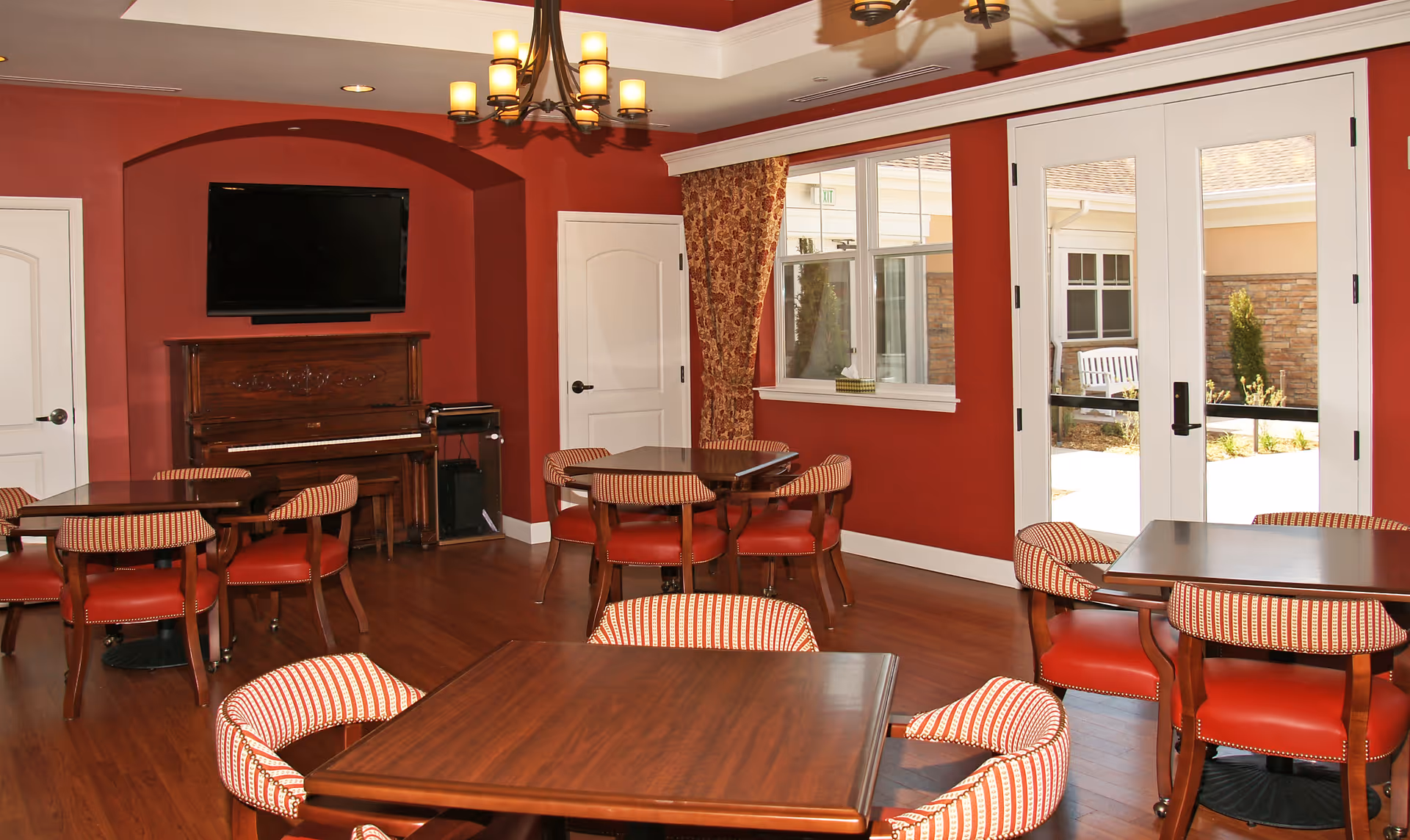 Interior view of a common area with red walls and wooden flooring. The room features several wooden tables surrounded by red and white striped cushioned chairs. A piano is placed against one wall beneath a mounted flat-screen TV. There are two white doors and a large window with patterned curtains. French doors lead to an outdoor patio area with a bench and plants visible outside.