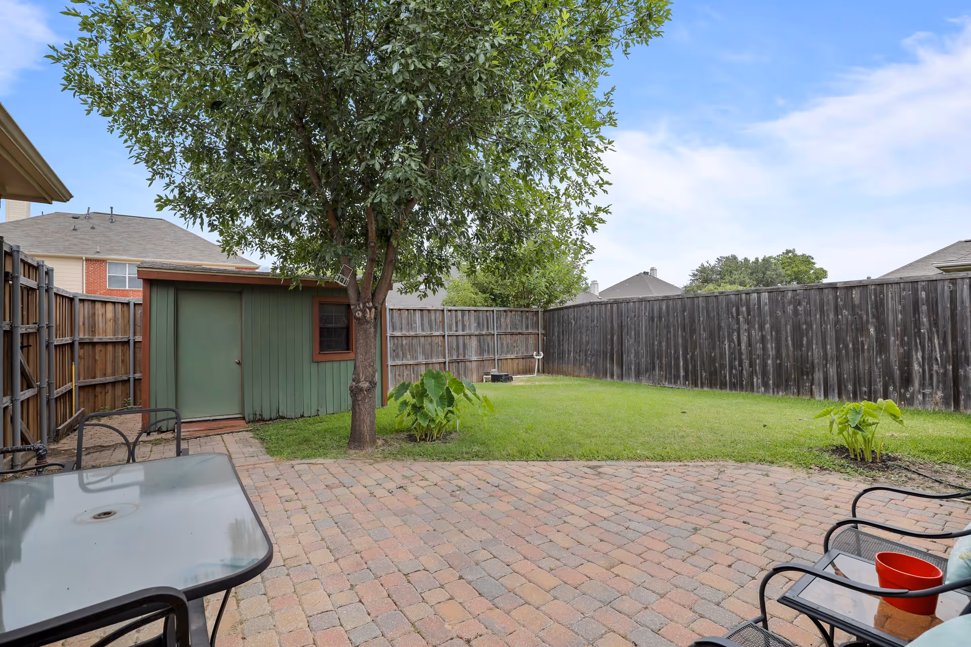 A backyard patio area with a brick-paved floor, metal chairs with cushions, a glass-top table, a green lawn, a tree, and a small green shed against a wooden fence under a partly cloudy sky.
