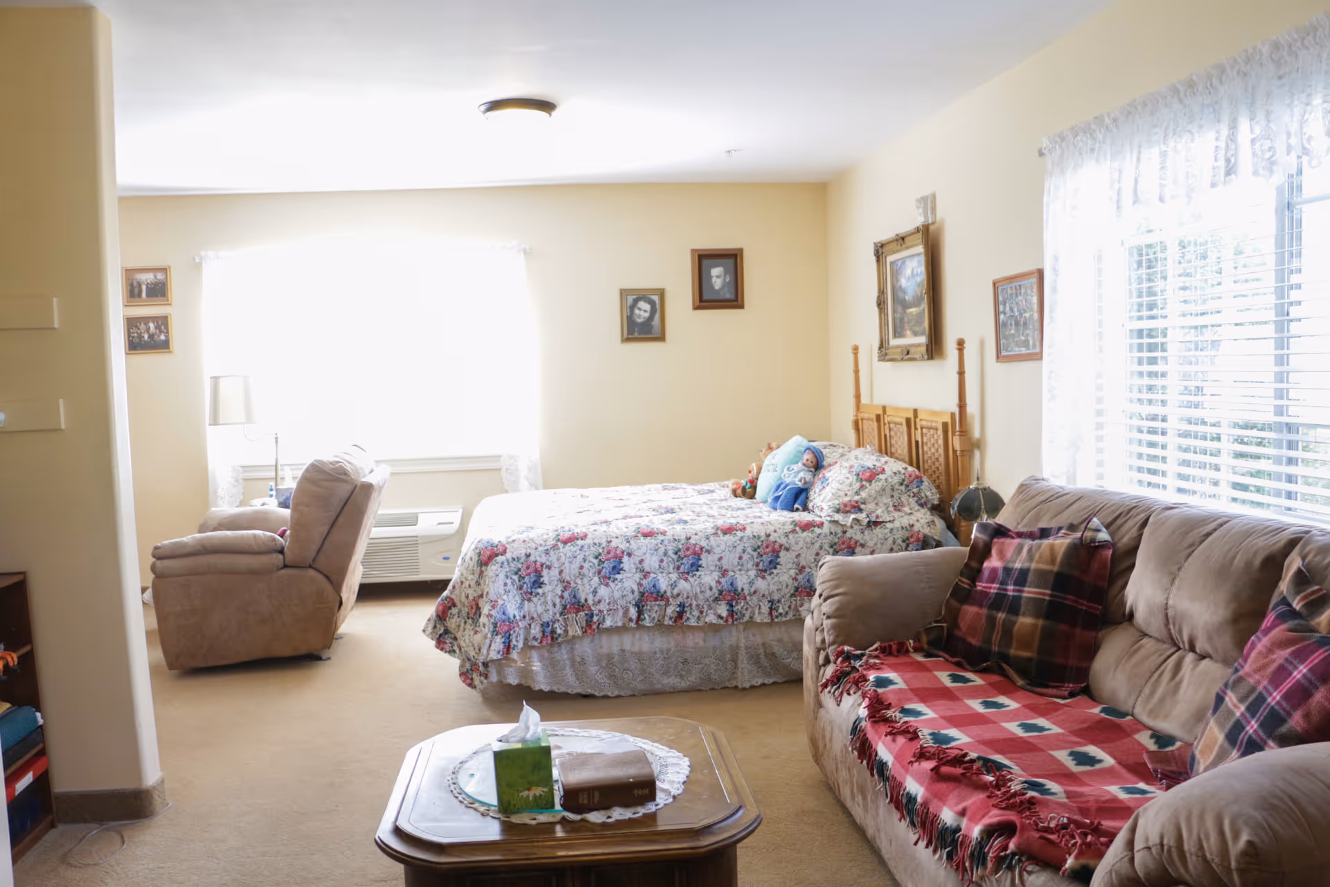 A cozy room with a bed covered in a floral quilt, a beige recliner chair near a window with white curtains, a beige sofa with plaid pillows and a red patterned throw, and a wooden coffee table with a tissue box and a book. The walls are light yellow and decorated with framed pictures and paintings.