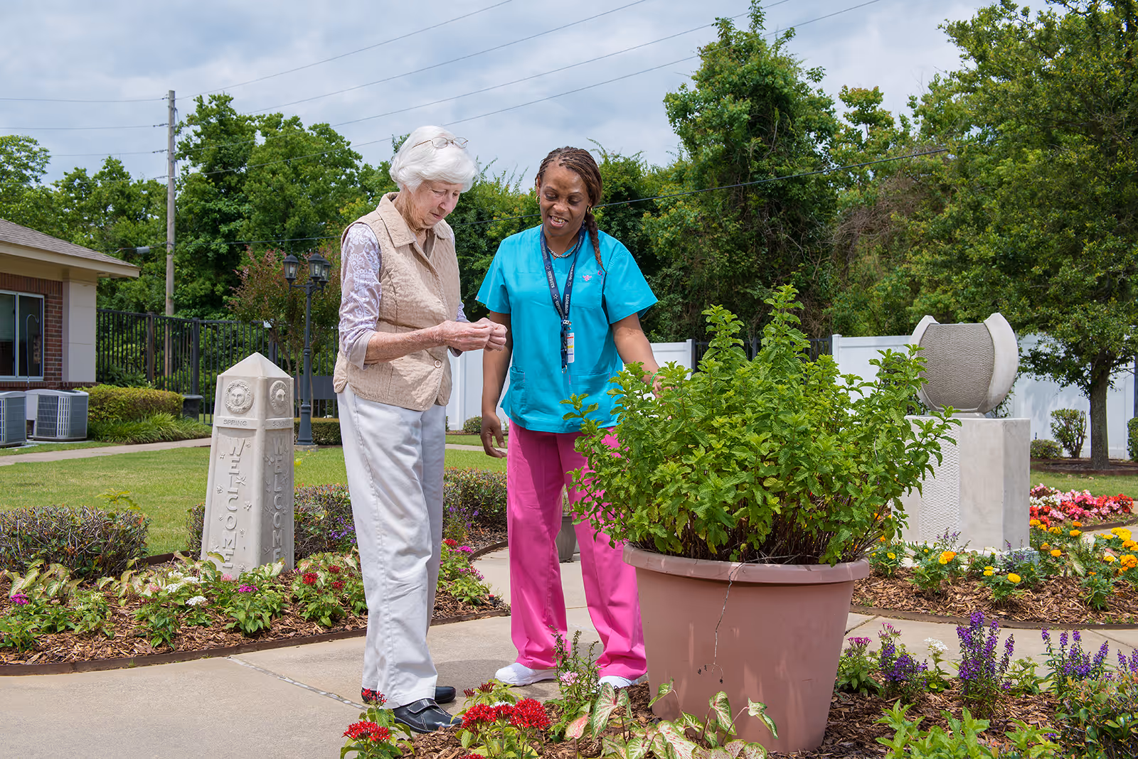 An elderly woman and a caregiver in teal scrubs stand together in a garden area of a retirement community. The elderly woman is looking at something in her hands while the caregiver smiles and looks on. The garden is well-maintained with various flowers, a large potted plant, and a stone monument with the word 'WELCOME' visible. Trees and a building are in the background under a cloudy sky.