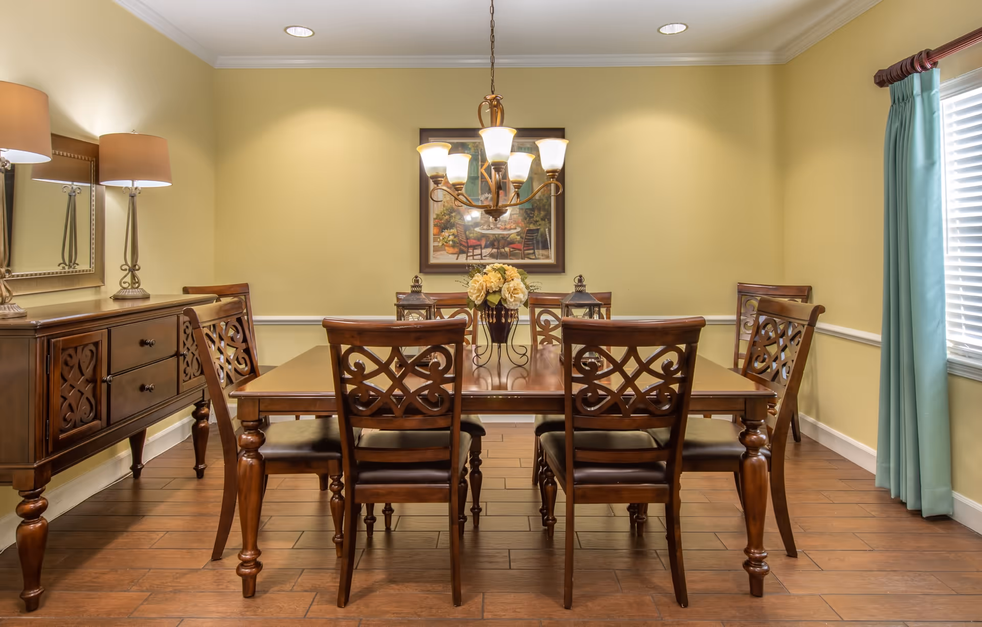 A dining room with a wooden dining table and six matching chairs. The table has a floral centerpiece and two lanterns. There is a sideboard with two lamps and a mirror on the left wall. A chandelier with multiple lights hangs above the table. The walls are painted light yellow, and there is a window with blue curtains on the right side. A framed painting hangs on the wall behind the table.