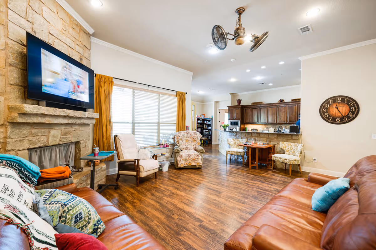 A spacious living room with wooden flooring, two brown leather sofas with colorful pillows, a stone fireplace with a mounted flat-screen TV, two armchairs near a large window with mustard yellow curtains, and a ceiling fan with lights. In the background, there is a kitchen area with wooden cabinets, a small dining table with two chairs, and a large wall clock.