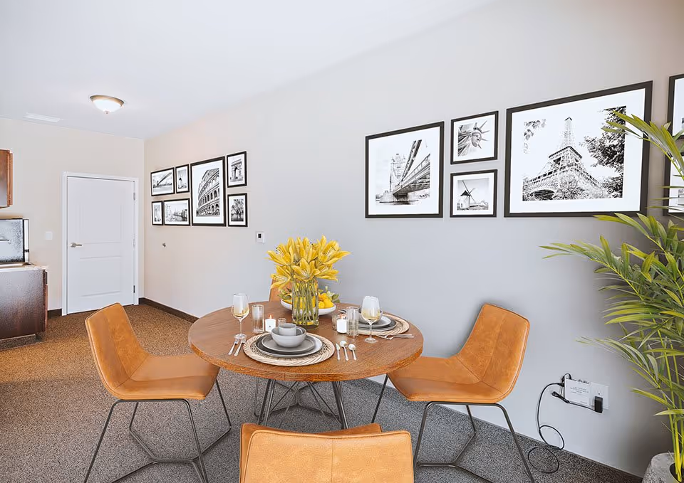 A dining area with a round wooden table set for four, featuring plates, bowls, glasses, and cutlery. A vase with yellow flowers and a bowl of lemons are on the table. Four brown leather chairs surround the table. The walls are decorated with black and white framed photographs of famous landmarks. A green plant is visible on the right side, and a kitchen area with wooden cabinets and a white door is in the background.