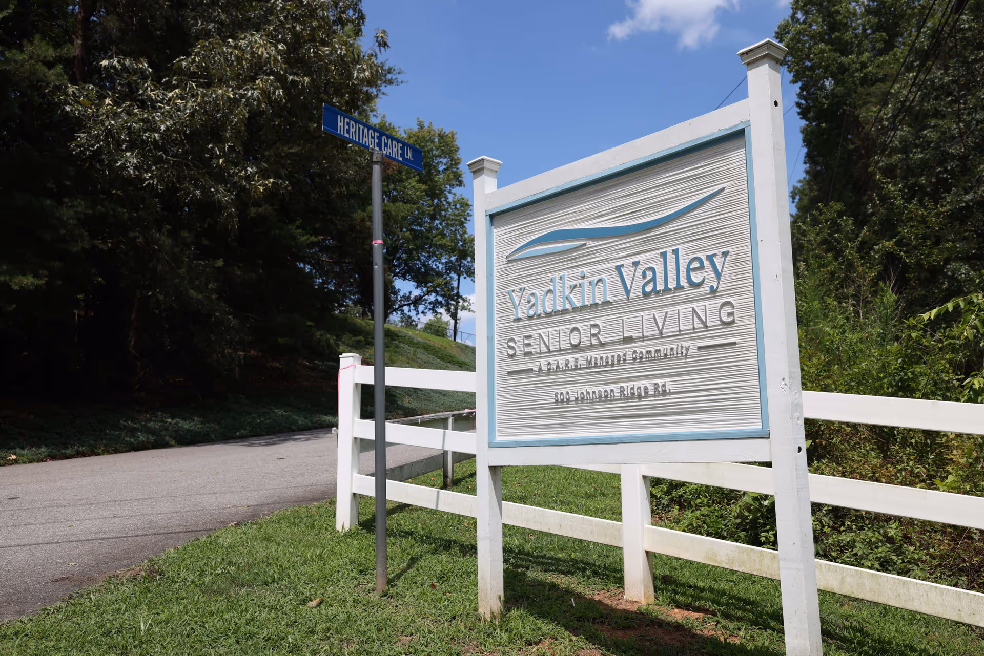 White wooden sign reading 'Yadkin Valley Senior Living' next to a road and white fence surrounded by trees.