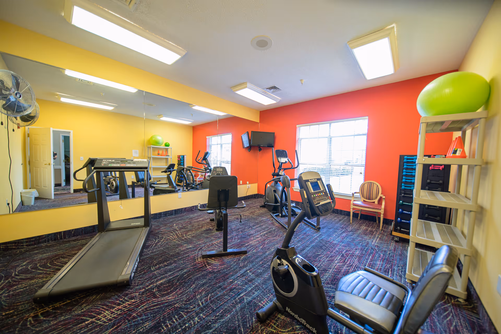 Bright exercise room with a treadmill, stationary bikes, mirrors, TV, and shelving with fitness equipment.