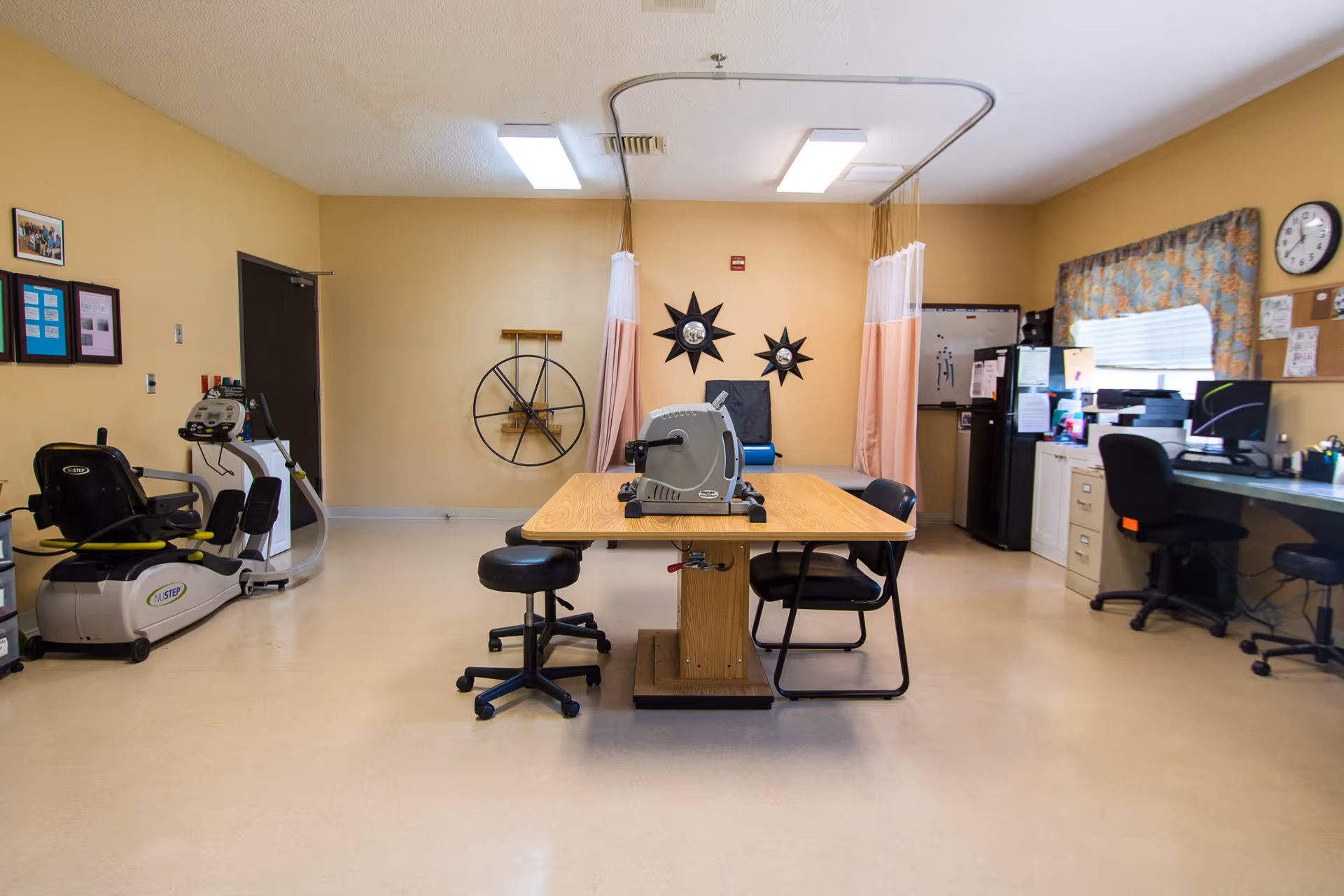 An interior activity/therapy room with a central table and chairs, exercise equipment on the left, desks and computers on the right, and privacy curtains at the back.