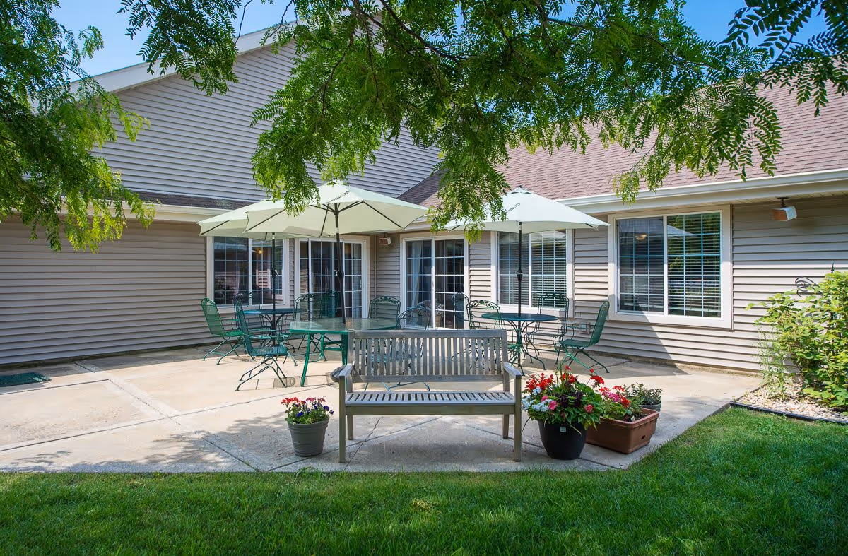 Outdoor patio area at a senior living facility with a wooden bench, several green metal tables and chairs, and two large white umbrellas providing shade. The patio is surrounded by a beige building with multiple windows and a green lawn with potted flowers in the foreground. Tree branches hang overhead, casting shadows on the patio.
