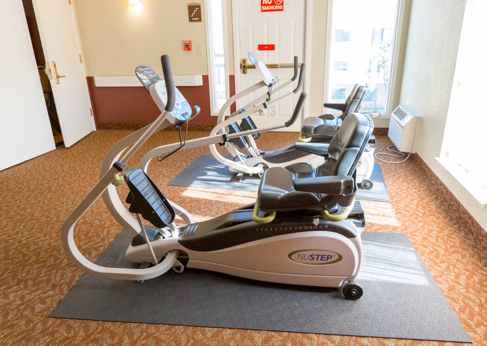 A small exercise room with two NuStep recumbent cross trainers placed on mats. The room has carpeted floors, a window letting in natural light, and a door with a no smoking sign above it.