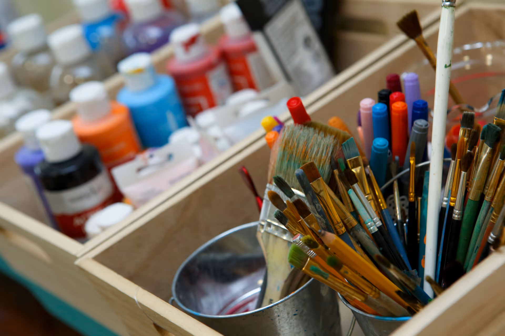 Close-up view of various paintbrushes and colorful paint bottles organized in wooden compartments, likely in an art or craft area.