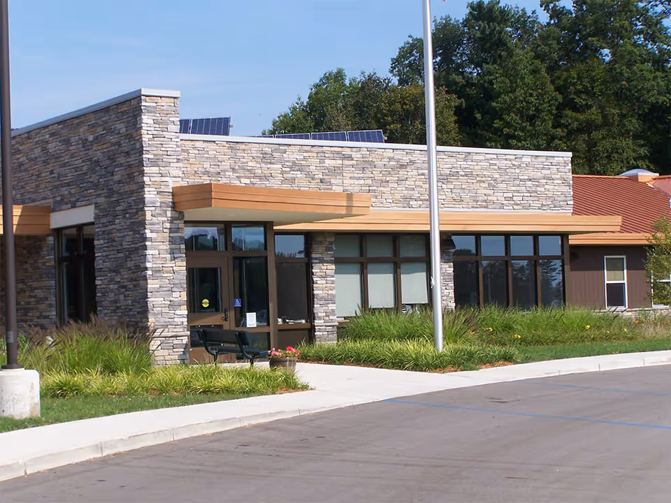 Exterior view of a modern building with stone and wood paneling, large windows, a flagpole, and landscaped greenery in front. The sky is clear and blue.