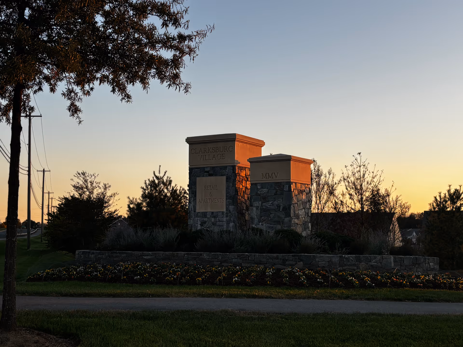 Stone monument sign at sunset with inscriptions 'Clarksburg Village', 'Retail & Apartments', and 'MMV', surrounded by landscaping and flowers, with trees and power lines in the background.