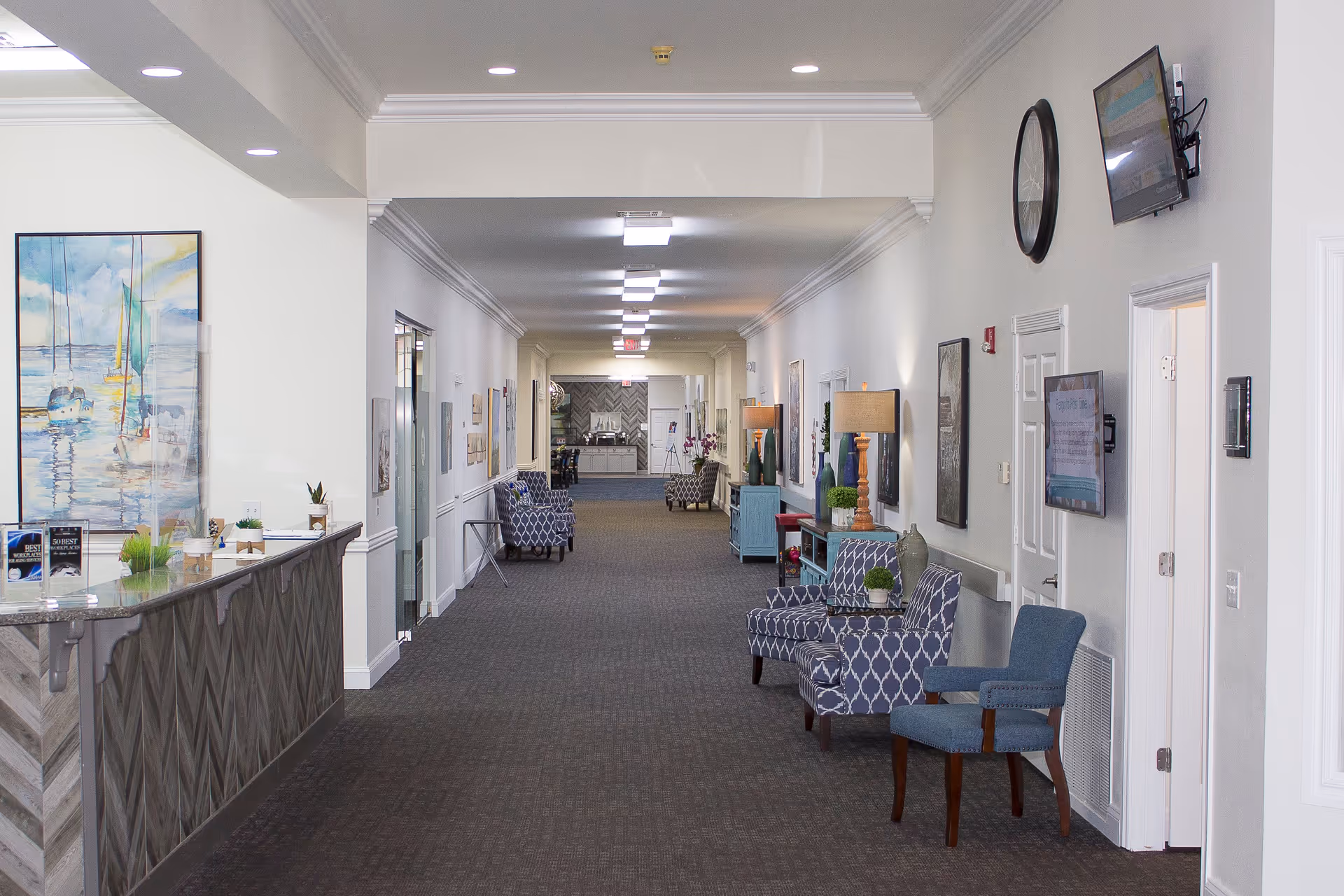 A long, well-lit hallway in a senior living facility with patterned armchairs and blue chairs along the walls. There are lamps, framed artwork, and flat-screen TVs mounted on the walls. A reception desk with decorative items and a large painting of sailboats is visible on the left side near the entrance.