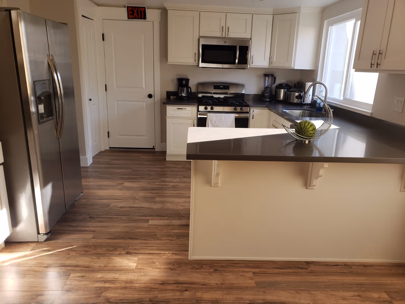 Sunlit modern kitchen with a peninsula countertop, stainless steel refrigerator and stove, white cabinets and wood-look flooring.