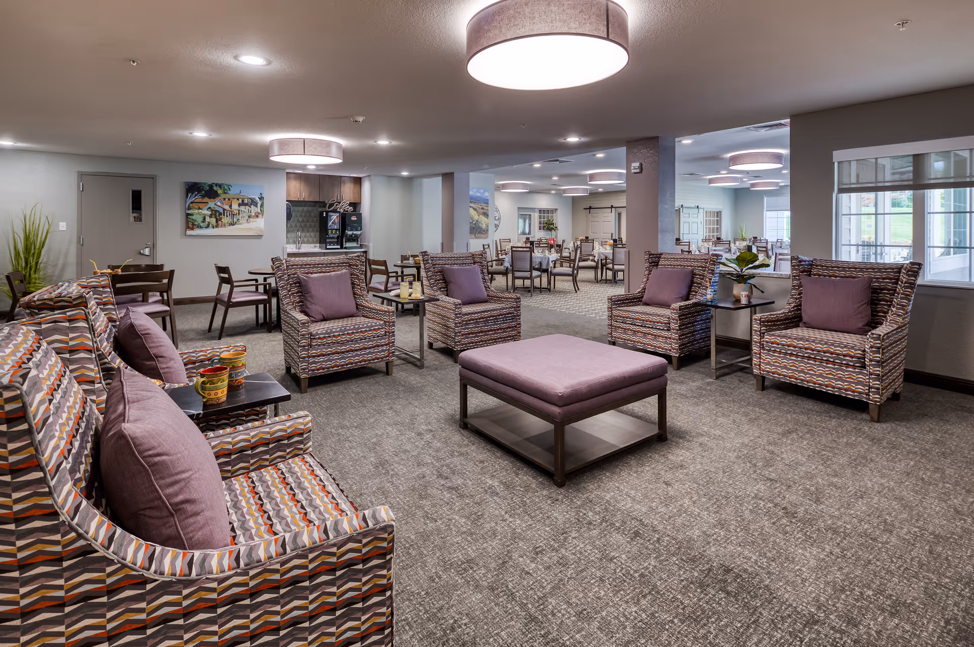 A spacious and well-lit common area in a senior living facility featuring patterned armchairs with purple cushions arranged around a central purple ottoman. In the background, there are dining tables and chairs, a coffee station, and large windows letting in natural light.
