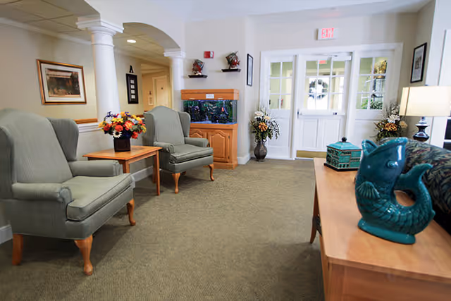 A cozy seating area in a senior living facility with two green upholstered armchairs, a wooden side table with a flower arrangement, an aquarium on a wooden cabinet, decorative vases with flowers, and a wooden console table with a teal ceramic fish sculpture. The space has carpeted flooring, white walls, and a glass door entrance in the background.