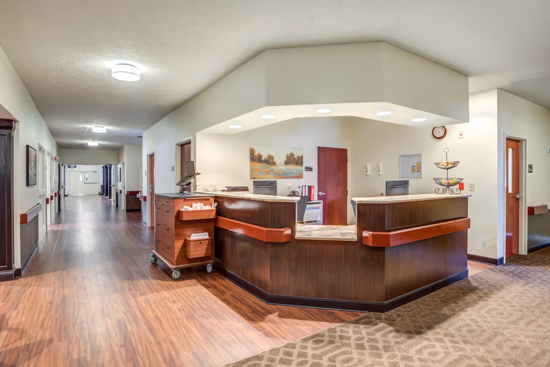 A clean and well-lit reception desk area in a senior living facility hallway. The desk is made of dark wood with a marble countertop and has a computer monitor, tissue box, and a tiered fruit basket on it. The hallway has wood flooring, beige walls, and several doors leading to other rooms. There is a painting on the wall behind the desk and a clock above it.