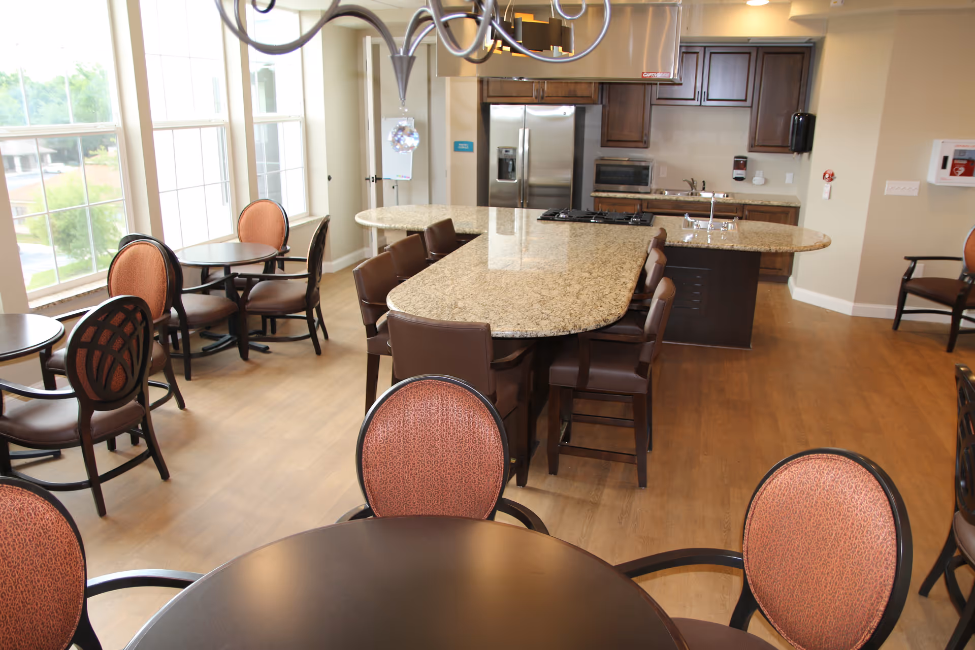 A bright dining area with multiple round tables and chairs with reddish upholstery. In the background, there is a kitchen area with a large granite island, bar stools, stainless steel refrigerator, microwave, and wooden cabinets. Large windows allow natural light to fill the room.