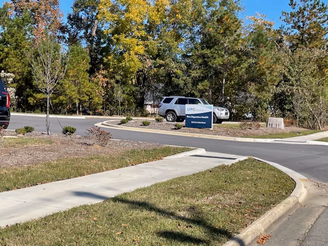 Curved driveway and sidewalk with a parked SUV, a blue sign, and trees in the background.