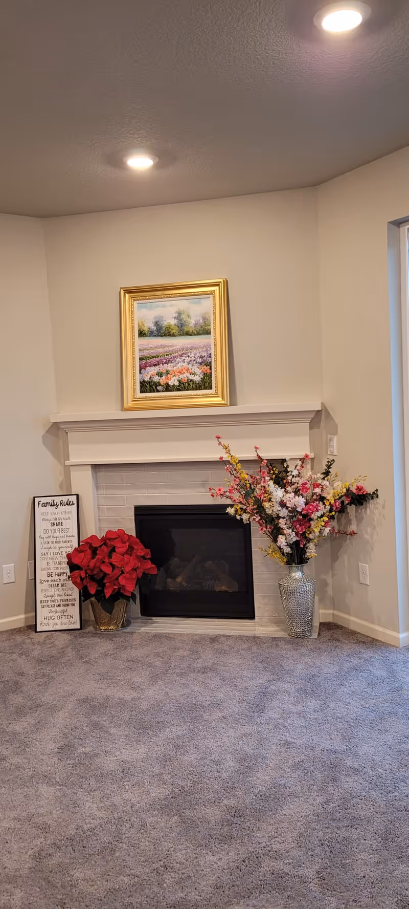 A cozy corner of a living room featuring a white fireplace with a framed floral painting above it. To the left of the fireplace is a red poinsettia plant and a decorative sign with family rules. To the right is a large vase filled with colorful artificial flowers. The room has beige walls, recessed ceiling lights, and a gray carpeted floor.