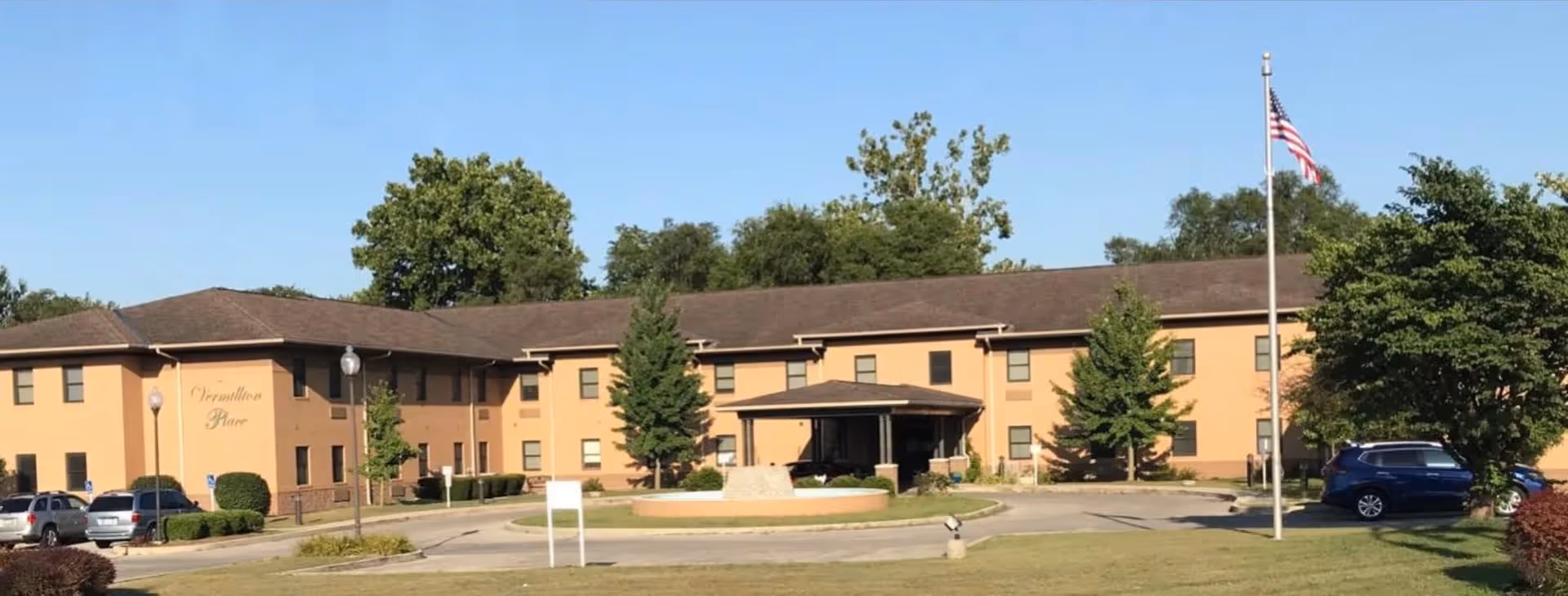 Front view of a two-story tan senior living building with a covered entrance, circular driveway, parked cars, and an American flag.
