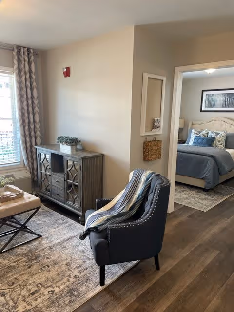 A cozy living area with a dark upholstered armchair draped with a striped blanket, a patterned area rug, a wooden console table with decorative items, and a window with patterned curtains. Through an open doorway, a bedroom is visible with a neatly made bed featuring blue and white pillows and a framed picture above the headboard.