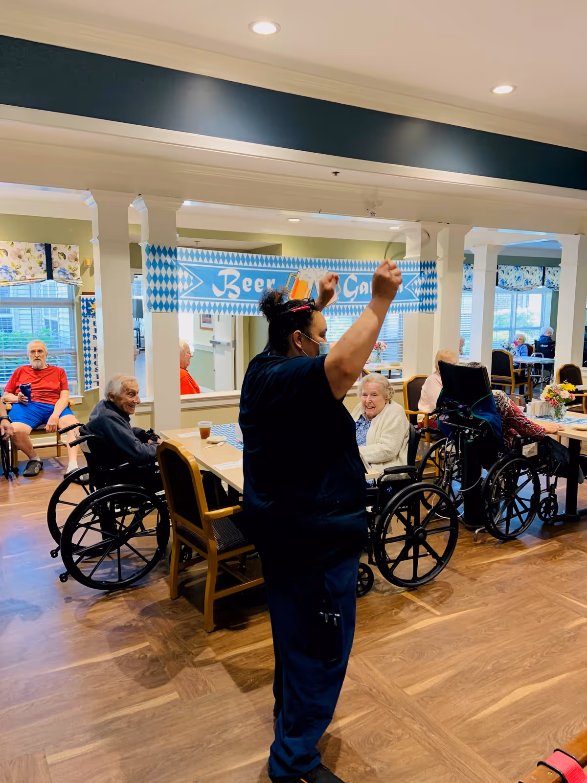 A staff member leads an activity while several residents in wheelchairs sit around tables in a bright communal dining/activity room.