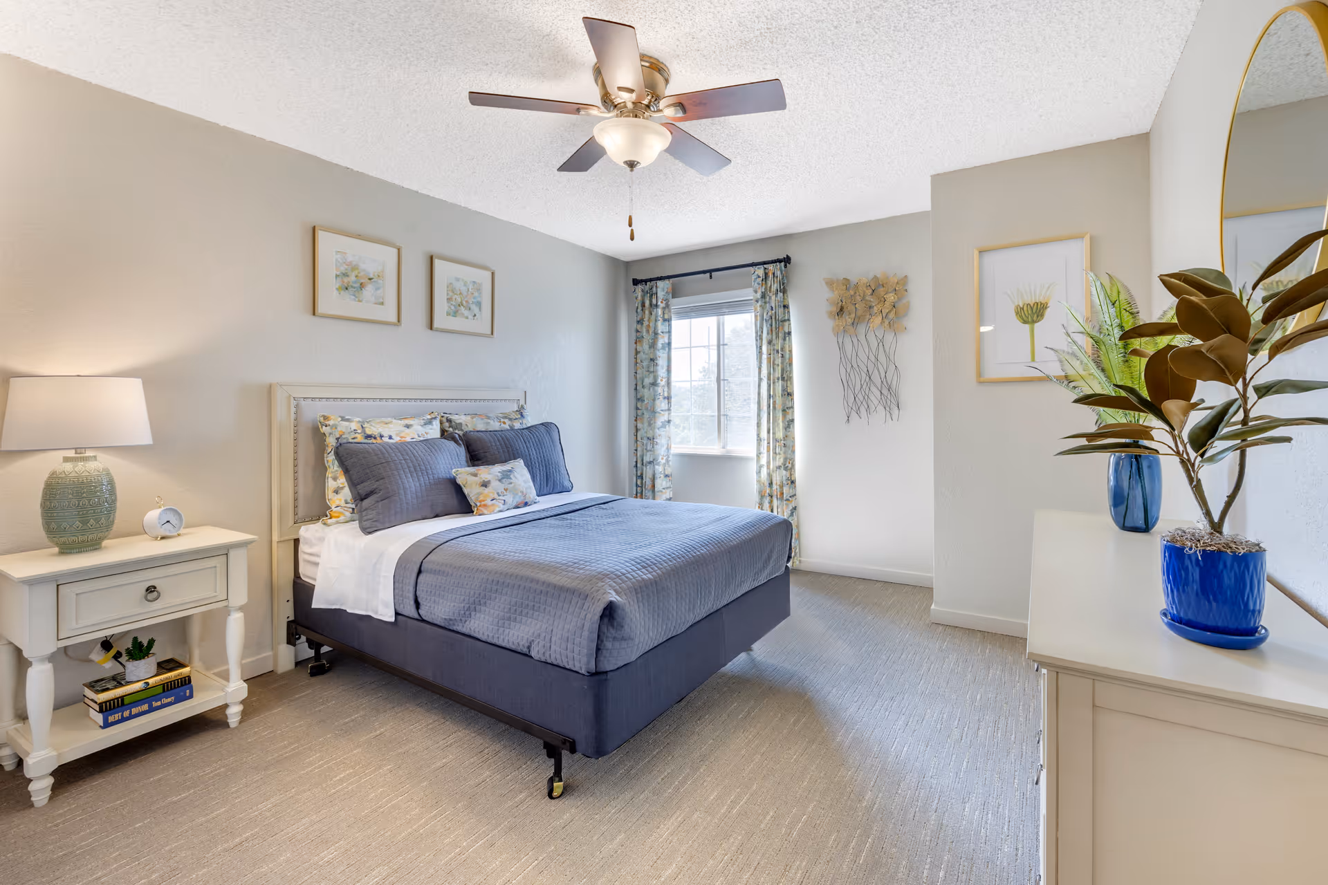 Bright, well-decorated bedroom with a made bed, bedside table and lamp, ceiling fan, window with curtains, and a dresser with plants.