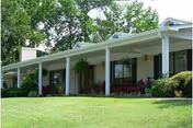 Single-story building with a long covered porch supported by white columns, surrounded by green grass and trees in the background.