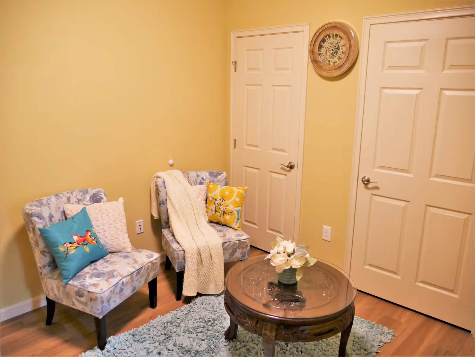 A cozy sitting area with two floral-patterned chairs, each adorned with decorative pillows and a cream-colored knitted throw blanket draped over one chair. A round wooden coffee table with a glass top holds a small vase of white flowers. The room has light yellow walls, two closed white doors, a round wooden wall clock, and a light blue shaggy rug on a wooden floor.