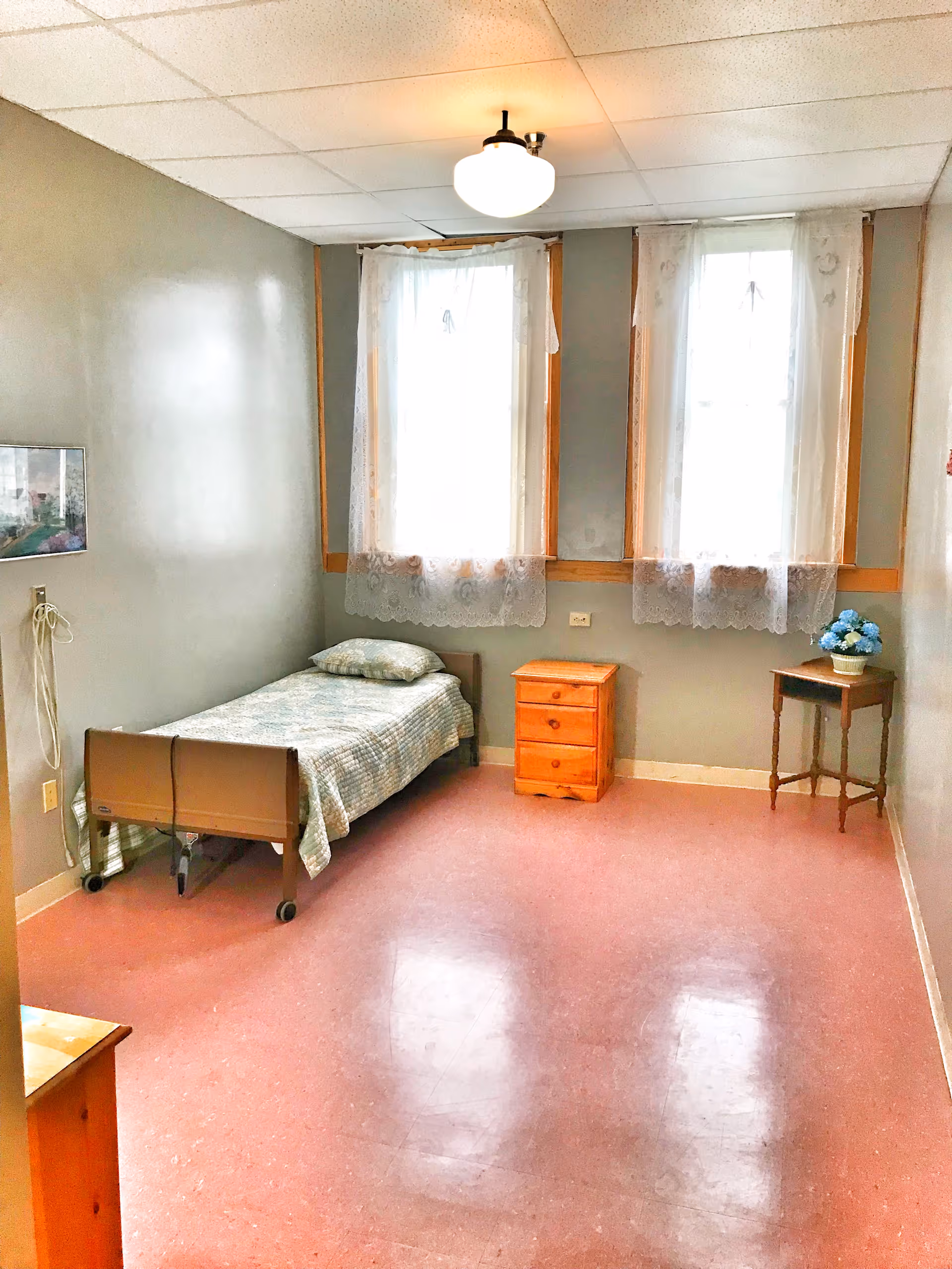 Small single-occupancy assisted living bedroom with a single bed, two wooden side tables, lace-curtained windows, and a pink linoleum floor.