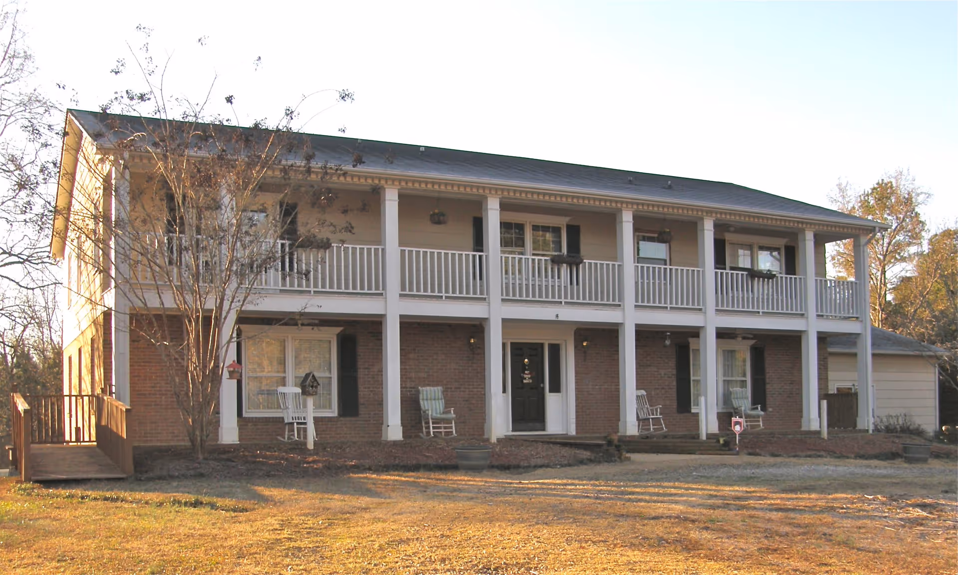 Two-story residential building with a brick lower level and beige upper level featuring a balcony with white railings. The front entrance has a black door flanked by windows with black shutters. There are rocking chairs on the porch and balcony, a tree without leaves in front, and a ramp on the left side leading to the porch.
