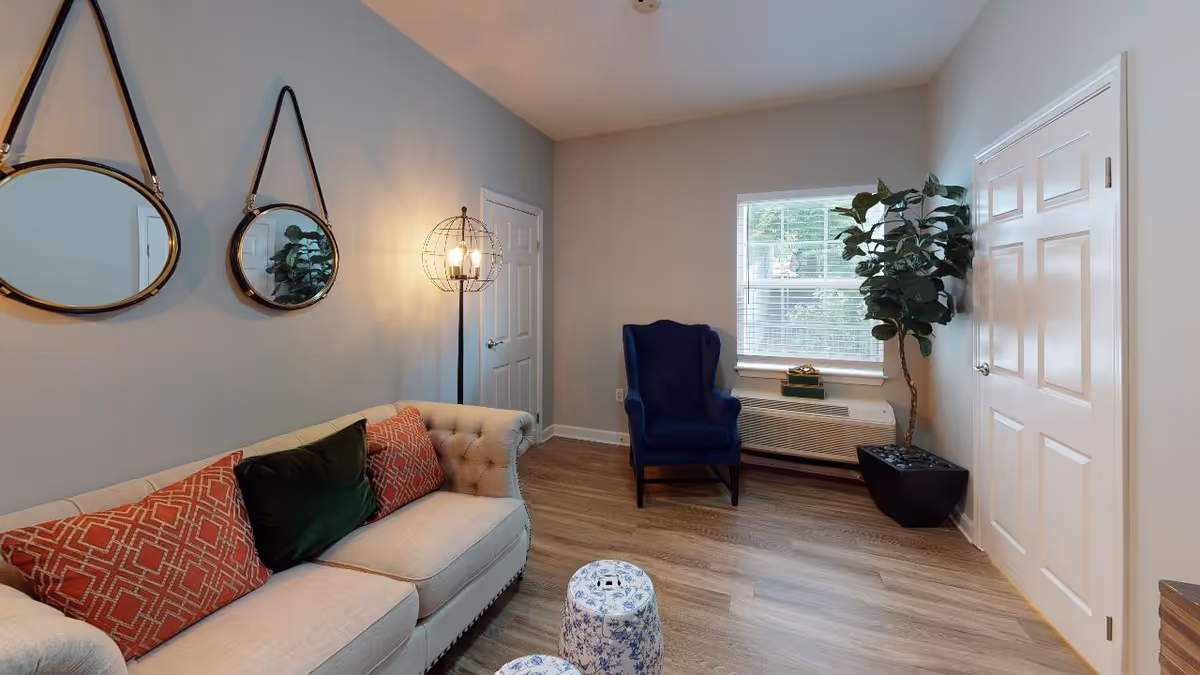 A cozy living room with a beige tufted sofa adorned with red and green patterned cushions, two round hanging mirrors on the wall above the sofa, a modern floor lamp with a spherical cage design, a blue armchair near a window with white blinds, a potted plant in a black square planter, and two small blue and white patterned stools on a wooden floor.