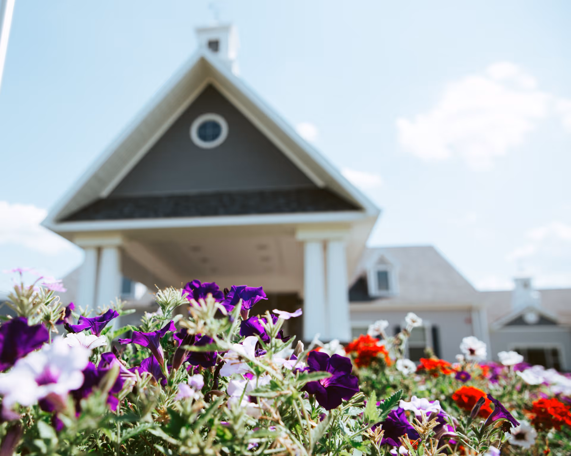 Colorful flowers in the foreground with the gabled entrance and columns of a senior living building in the background.