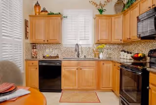 A bright kitchen with light wood cabinets, a windowed sink, black appliances, a mosaic tile backsplash, and a small dining table with place settings.