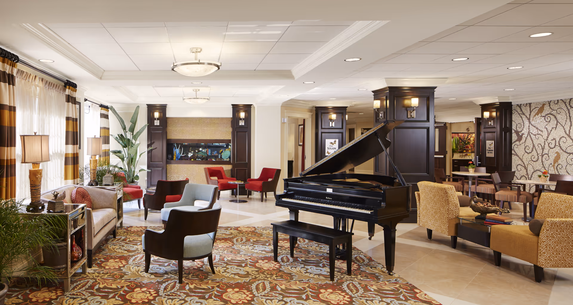 Well-lit communal lounge with a grand piano at center surrounded by upholstered chairs, sofas, plants, and a patterned rug.