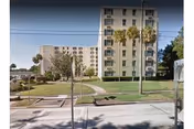 Multi-story beige apartment building with palm trees, lawn, and a sidewalk along the street.