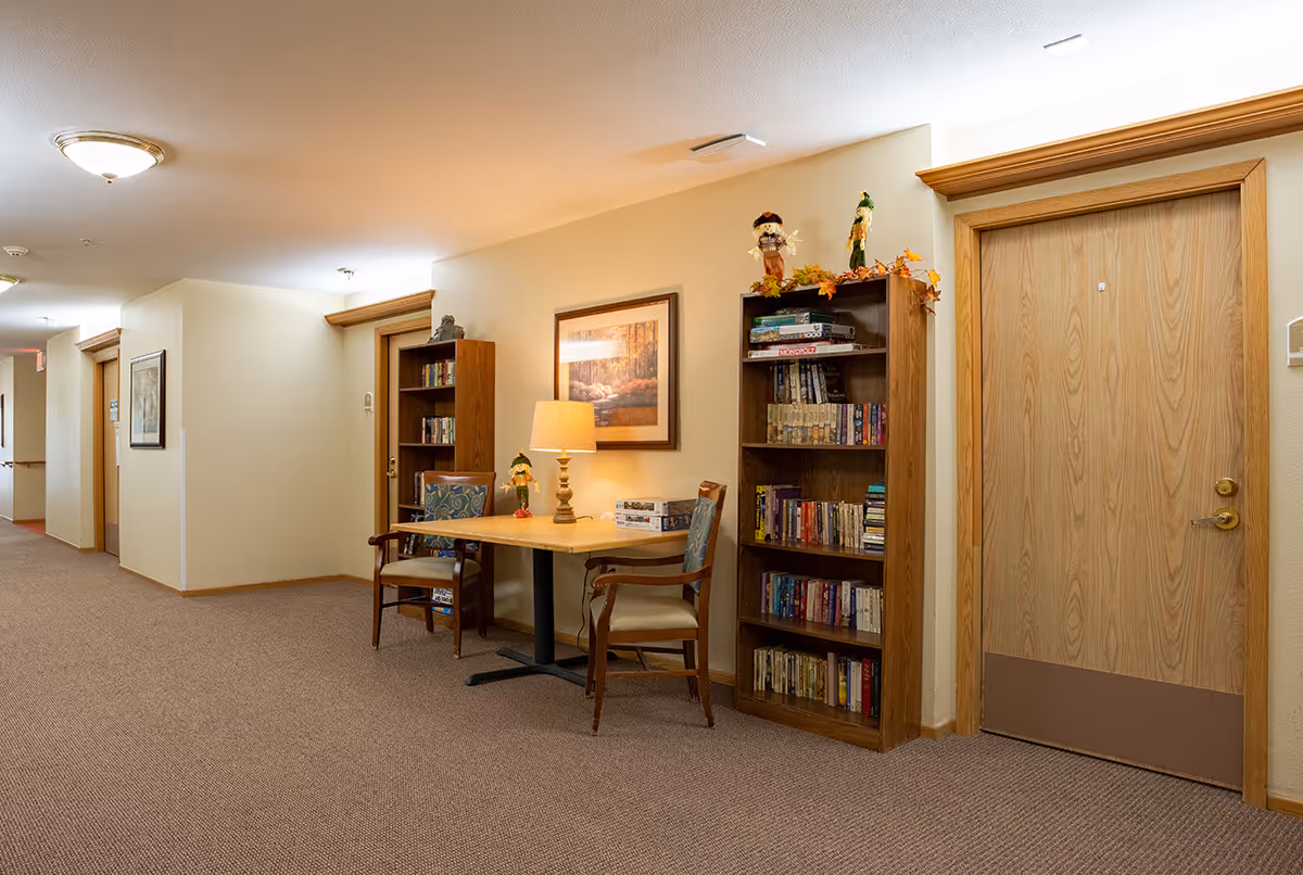 A well-lit hallway in an assisted living facility with beige walls and carpeted floor. There are wooden doors along the hallway, two wooden bookshelves filled with books, a small table with a lamp, two chairs, and autumn-themed decorations on top of the bookshelf. A framed picture hangs on the wall above the table.