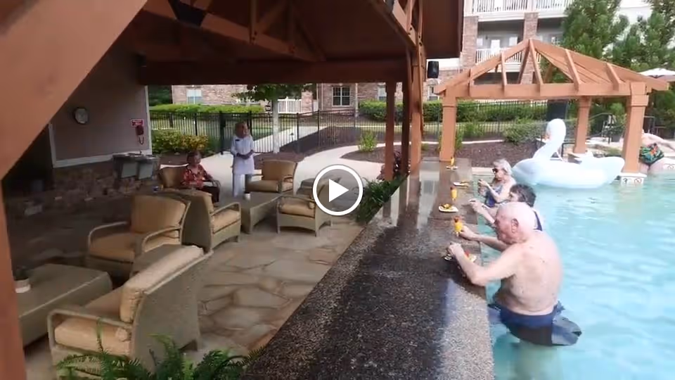 Seniors sitting in a pool at a swim-up bar with lounge seating under a wooden pergola at an outdoor pool.