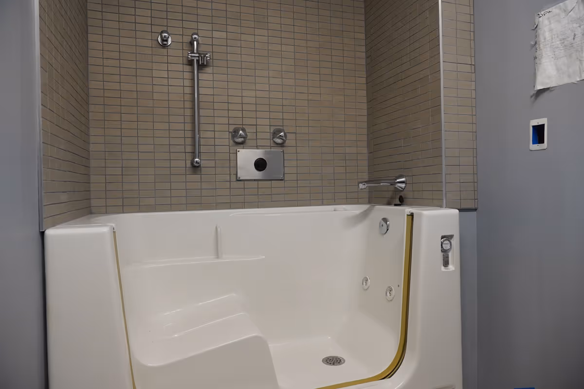 White walk-in bathtub with built-in seat and jets set against a beige tiled shower wall with faucet and grab bar.
