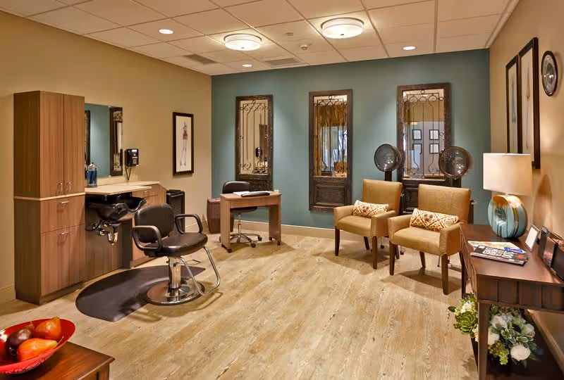 Interior view of a salon area in a senior living facility with a hair washing station, salon chair, two beige armchairs with patterned pillows, a small desk with a chair, decorative mirrors on a teal accent wall, a side table with a lamp, and framed artwork on the walls.