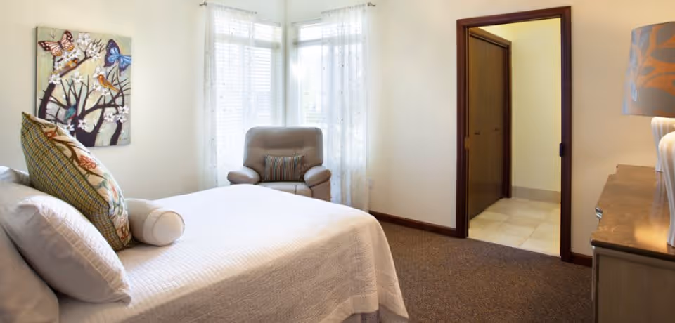 A cozy bedroom in a senior living facility featuring a neatly made bed with multiple pillows, a comfortable armchair near a window with sheer curtains, a dresser with a lamp, and a doorway leading to another room.
