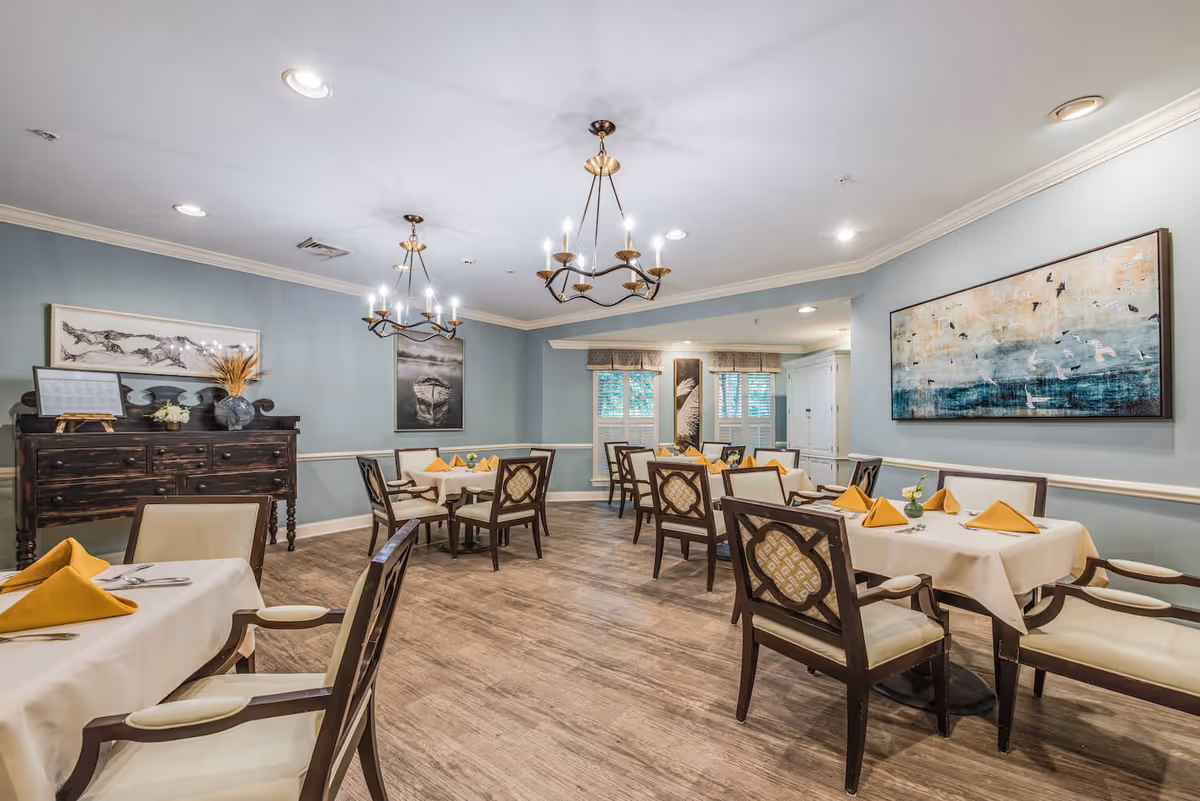 A bright dining room with multiple set tables and chairs, yellow napkins, chandeliers, and wall artwork.