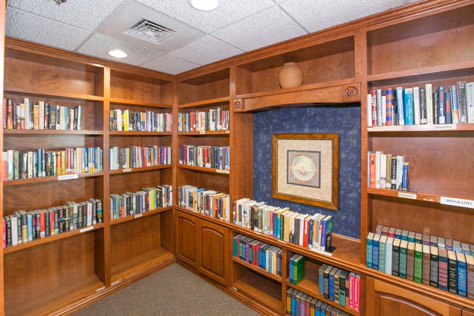 Interior view of a library room with wooden bookshelves filled with books. The shelves are arranged in an L-shape, and there is a framed picture hanging on a blue wall section in the middle of the shelves. The ceiling has recessed lighting and a vent.
