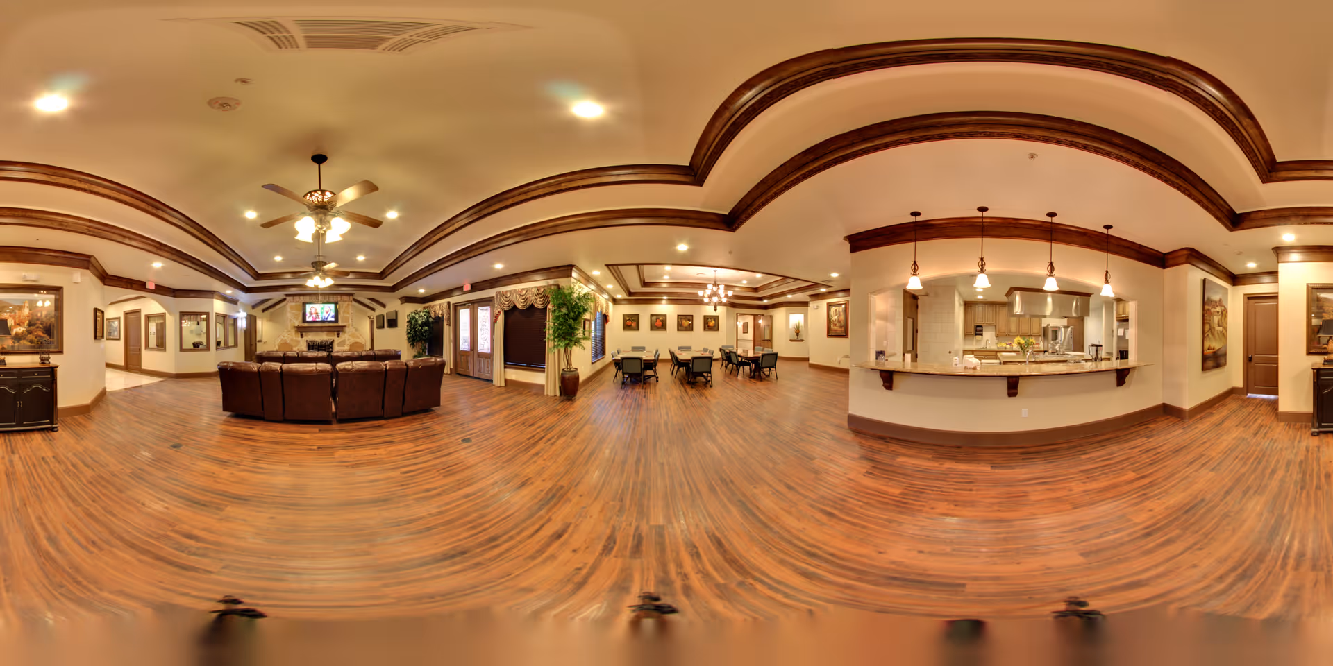 Panoramic interior of a senior living facility common area showing a seating lounge with a TV, dining tables, and a serving counter/kitchen.