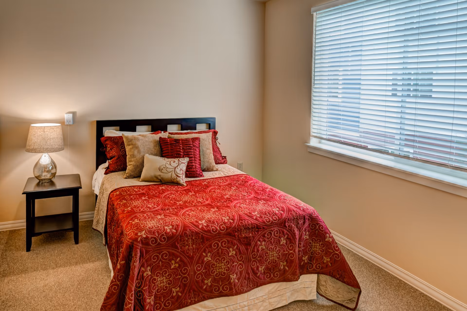 A cozy bedroom with a bed covered in a red patterned bedspread and multiple decorative pillows. There is a small black nightstand with a lamp on it next to the bed. A large window with white blinds is on the right wall, allowing natural light into the room.
