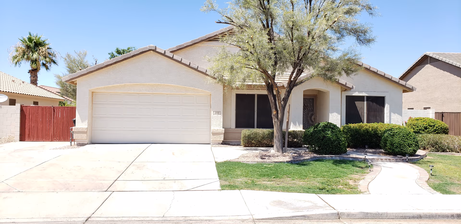 Single-story stucco home with a two-car garage, front yard, tree, and trimmed shrubs under a clear blue sky.
