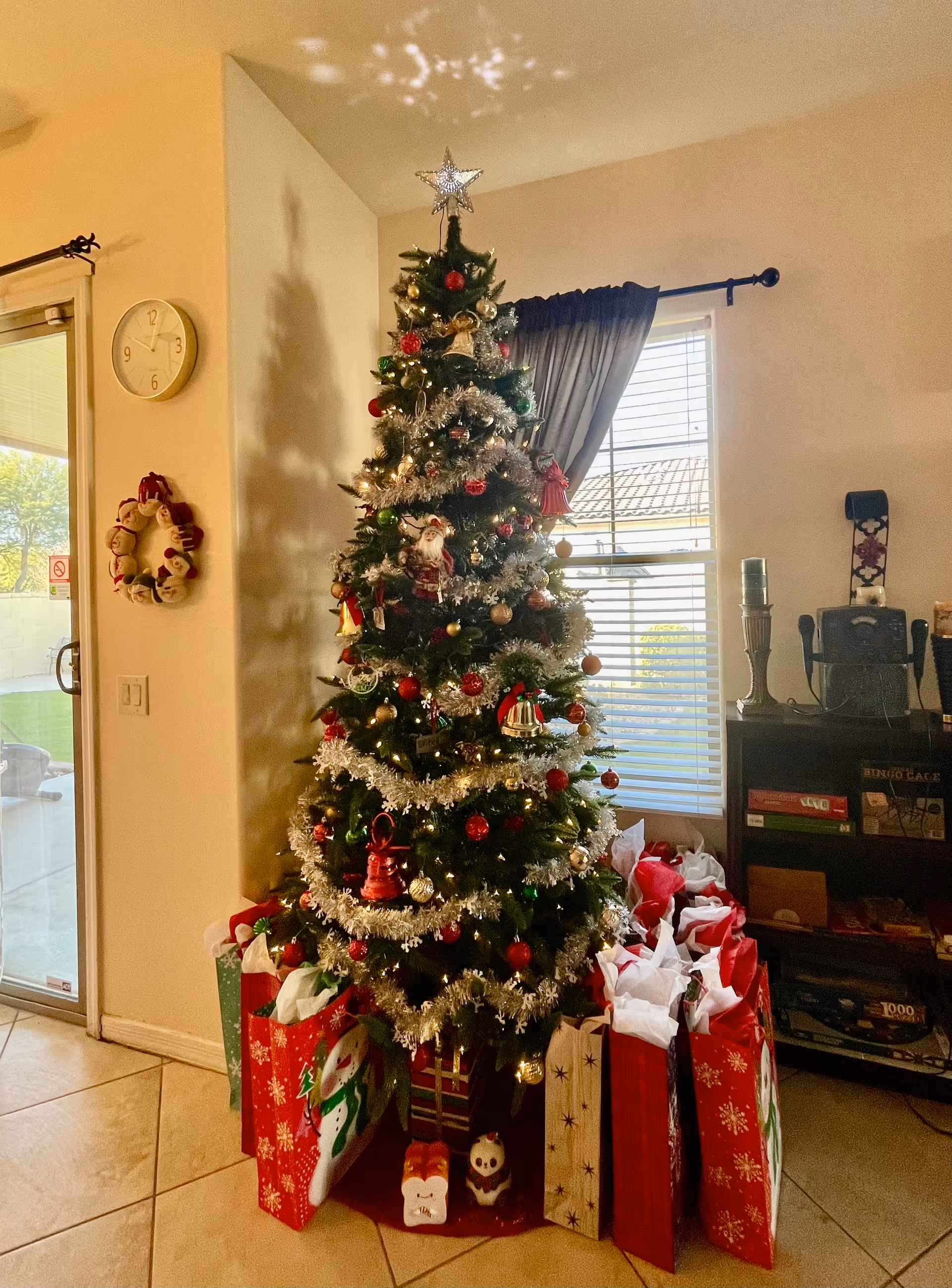 A decorated Christmas tree with a star on top, adorned with ornaments and tinsel, standing in a corner of a room. Around the base of the tree are numerous gift bags filled with tissue paper. To the left, there is a wall clock and a holiday wreath hanging on the wall near a sliding glass door. To the right, a window with blinds and a dark curtain is visible, along with a shelf holding various items including board games and candles.